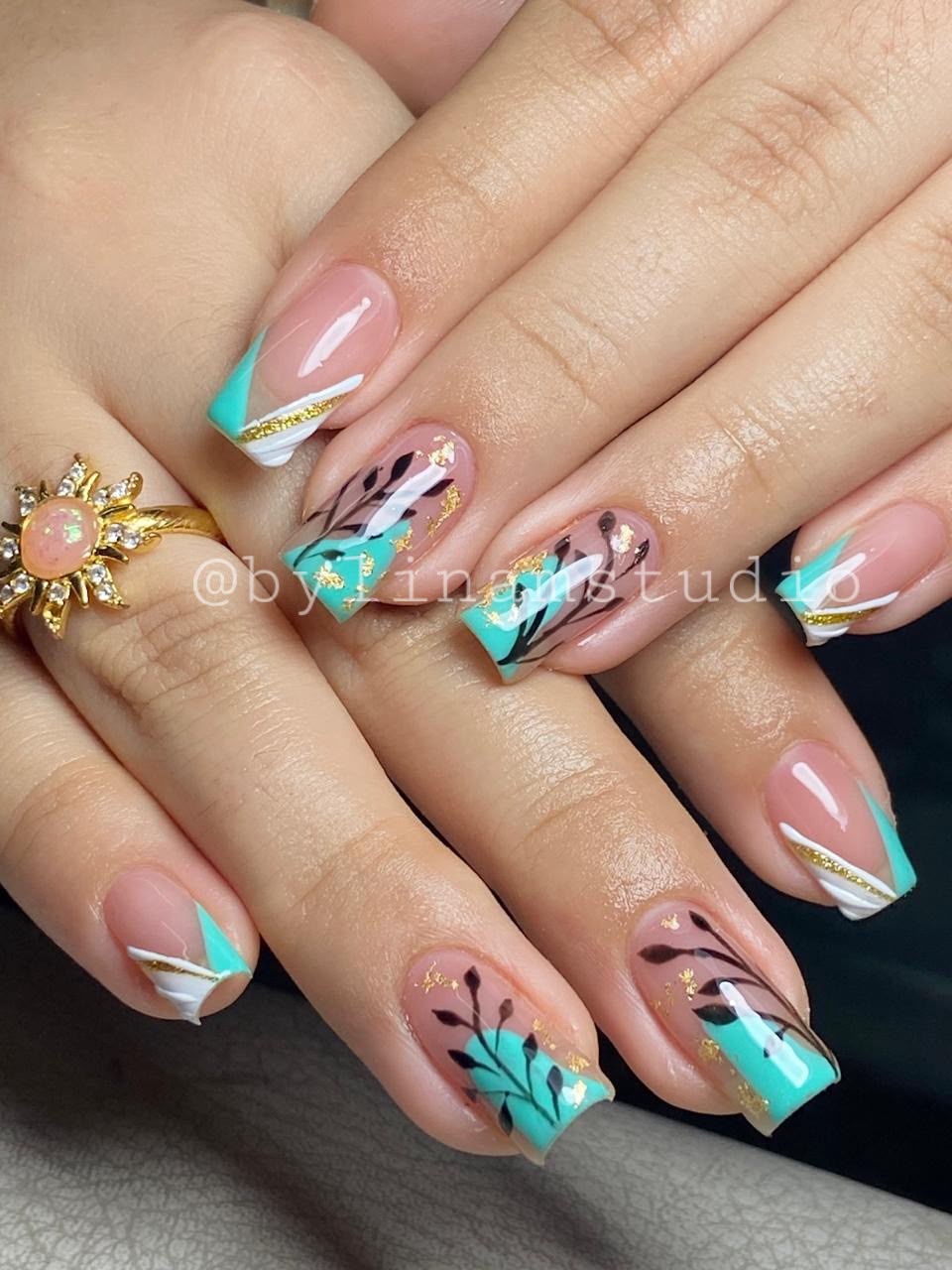 Manicura cuadrada nude con bloques turquesa, líneas doradas y hojas en negro
